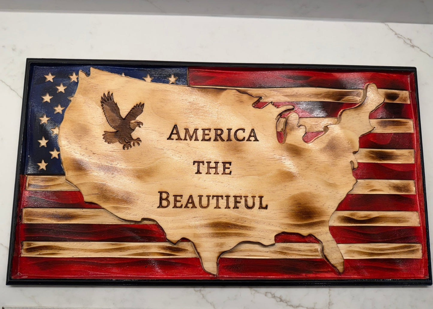 Wooden plaque with American flag design, eagle, and 'America the Beautiful' text on a marble surface.